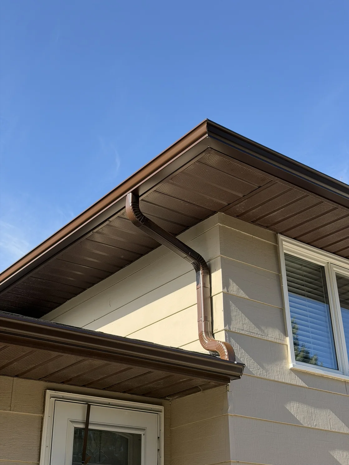Brown downspout with elbows connecting an upper gutter to a lower gutter on a Regina home.